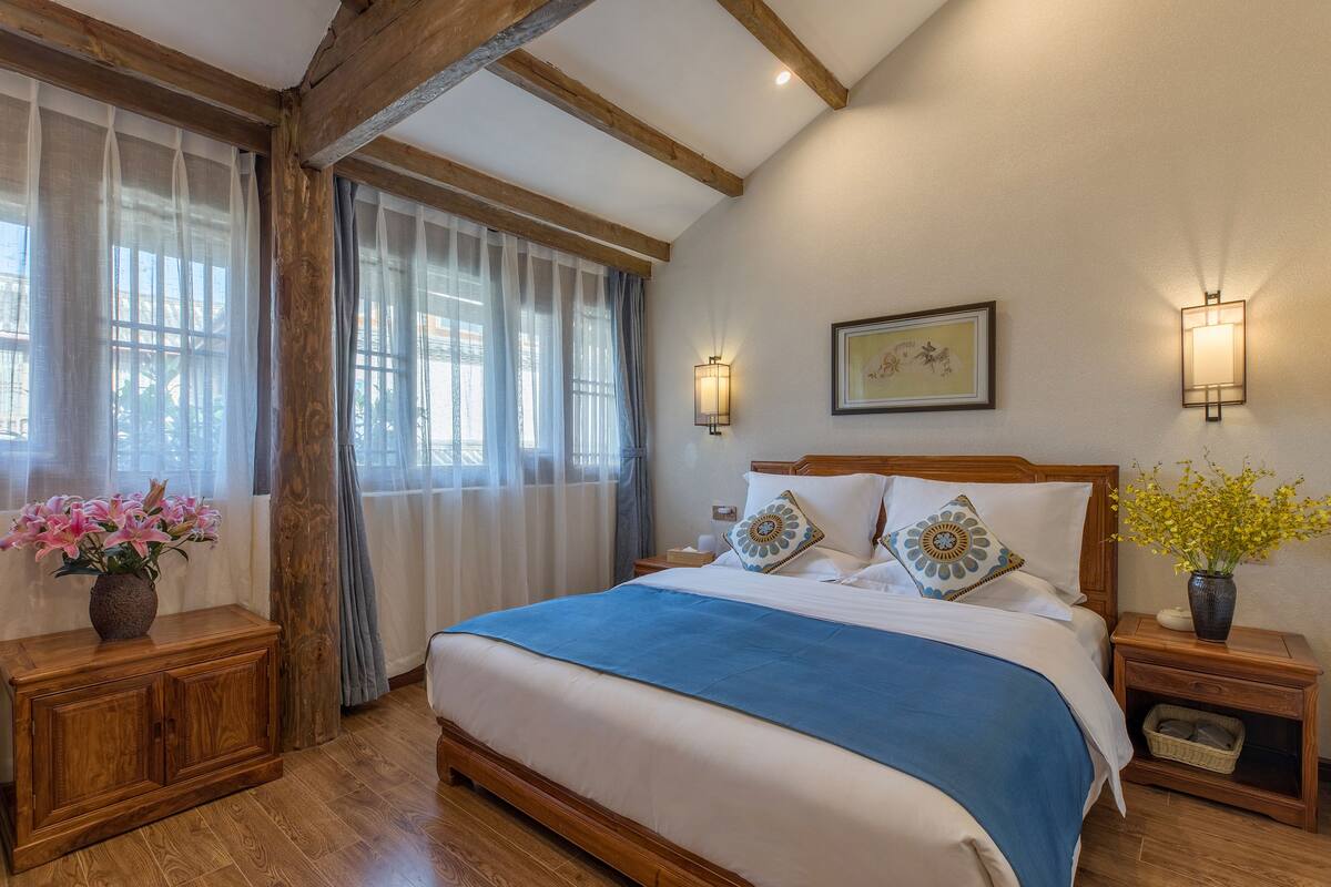Een slaapkamer met een houten bedframe, een blauw bedlaken, twee nachtkastjes met lampen, een bijzettafeltje met een bloevas en een raam met dunne gordijnen.