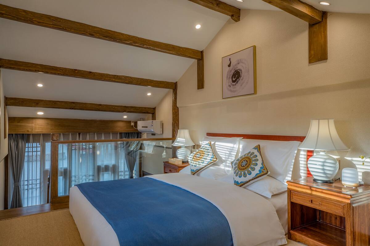 Een slaapkamer met een groot bed, houten balken, een raam met gordijnen en een nachtkastje met lampen.