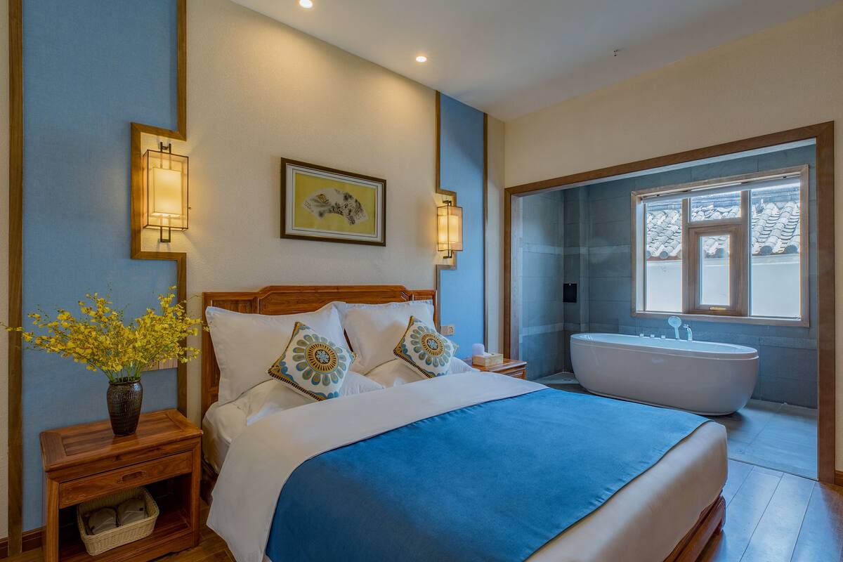 Een slaapkamer met een groot bed, een nachtkastje met een vaas bloemen, een wandlamp en een bad in de aangrenzende badkamer.