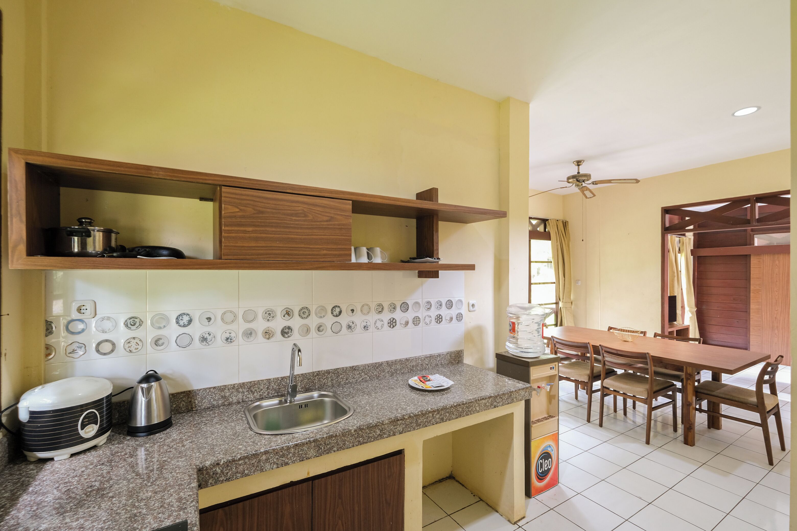 A kitchen with granite countertops, a sink, and wooden cabinets. There is a dining table with chairs and a ceiling fan.