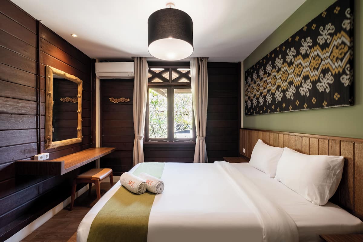 Ein Hotelzimmer mit Bett, einer Holzbank, einem Schreibtisch, einem Spiegel und einem Fenster mit Vorhängen.