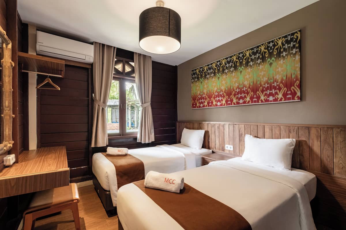 Ein Hotelzimmer mit zwei Betten, einem Schreibtisch, einem Kleiderhaken und einem großen abstrakten Gemälde an der Wand.