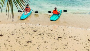Two kayaks on a sandy beach with clear blue water and a palm tree in the foreground. - Mutiara Carita Cottages (Carita)