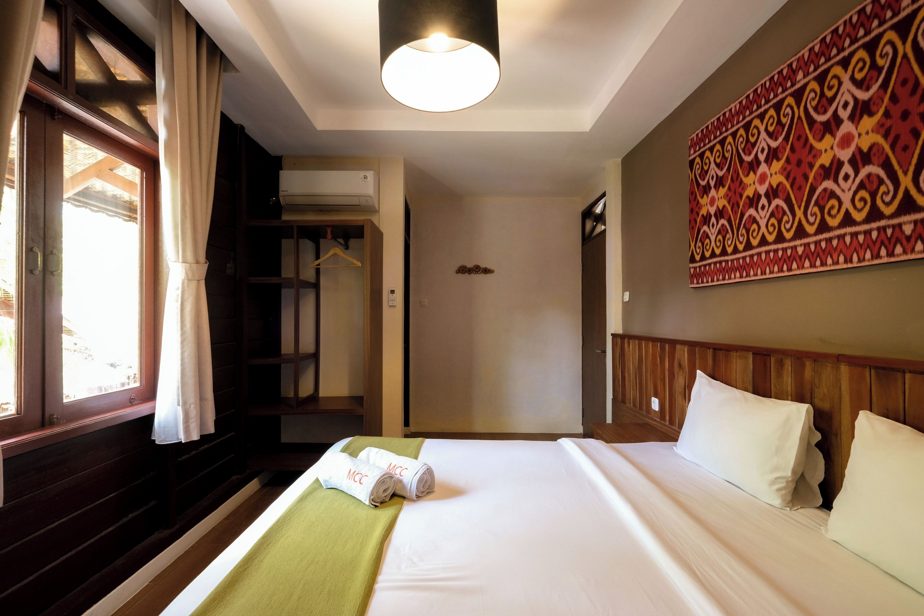 A hotel room with a bed, a green throw blanket, a hanging coat rack, and a large patterned wall hanging.