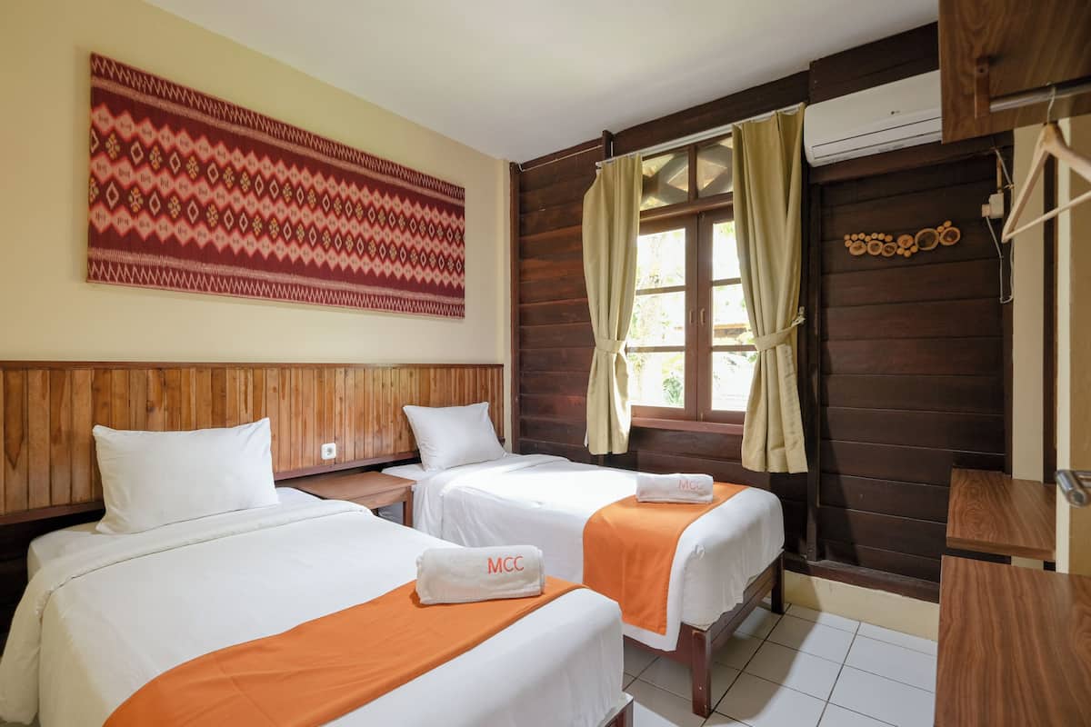 Ein Hotelzimmer mit zwei Betten, Holz-Kopfteilen, einem großen Wandteppich mit Muster sowie einem Fenster mit Vorhängen und einer Klimaanlage.