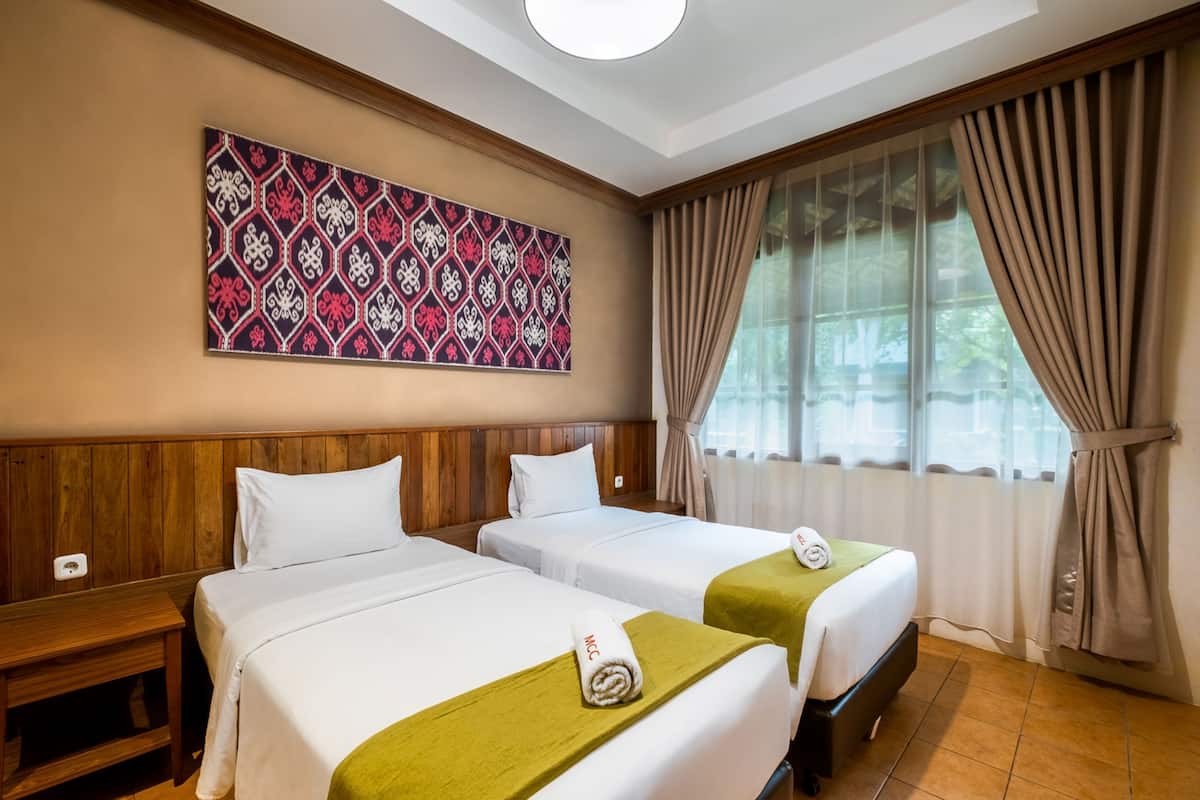 Ein Hotelzimmer mit zwei Betten, einem hölzernen Kopfteil, einem großen Fenster mit Vorhängen und einem Wandbehang mit Muster.