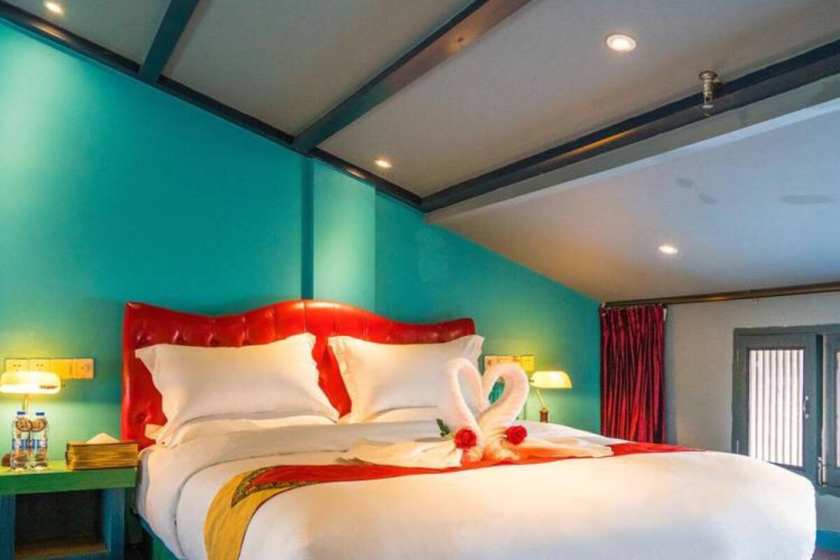 A large bed with a red headboard, white pillows, and a decorative swan made of towels. There are two bedside tables with lamps and a window with curtains.