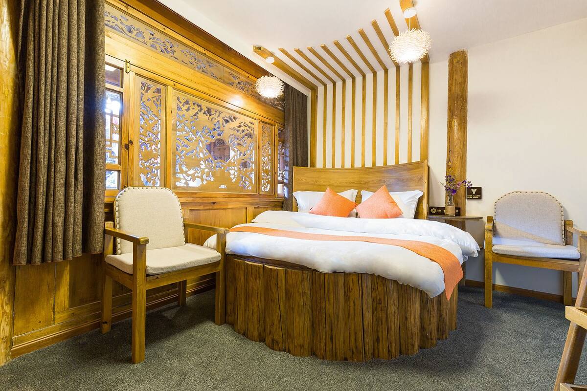 Een bed met houten frame en een rond houten voeteneinde, twee stoelen en een decoratief paneel aan de muur.