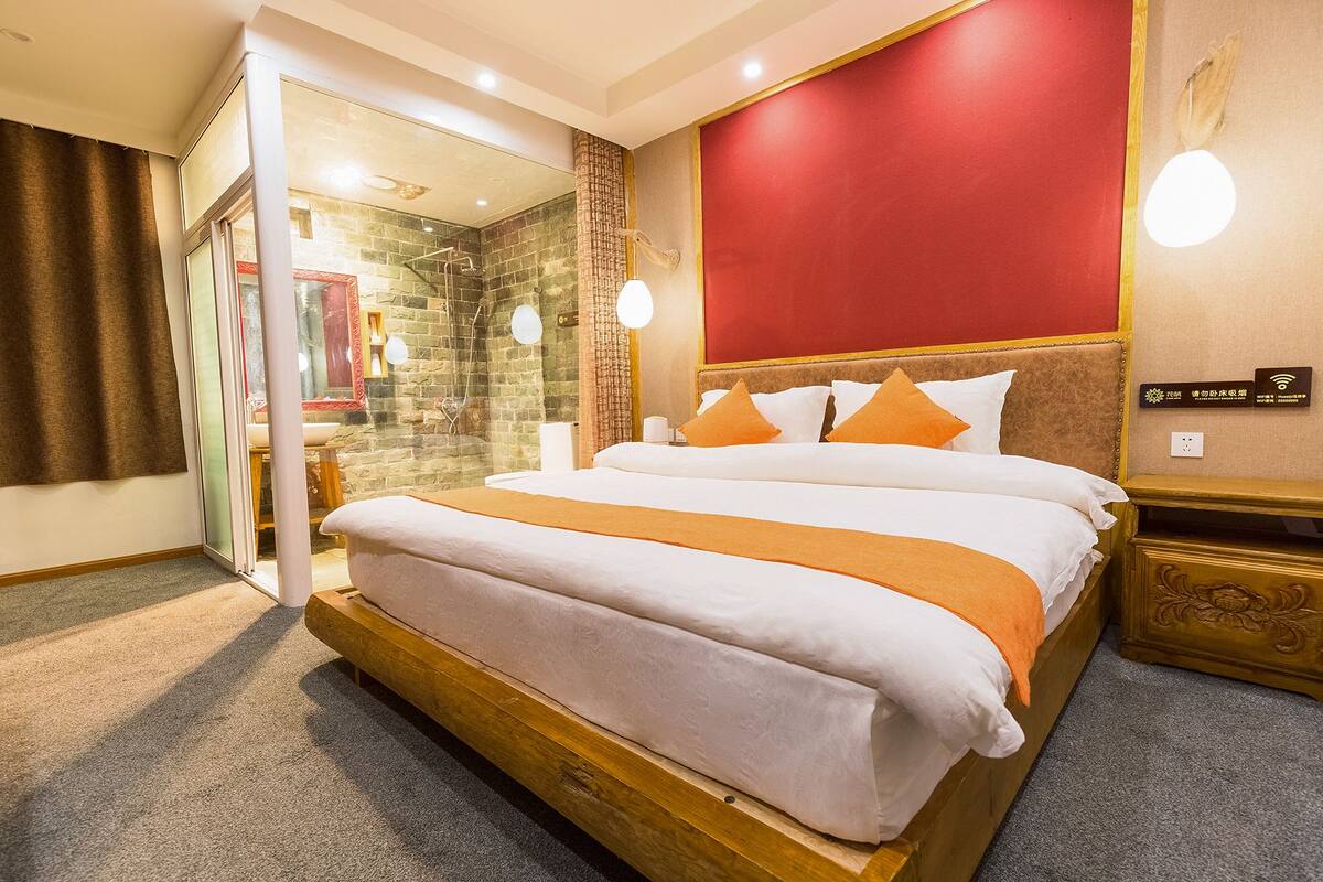 Hotelkamer met groot bed, rode accentmuur, houten hoofdeinde en douchecel van glas.