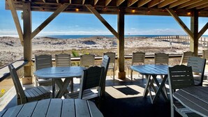 Ett utomhusområde med sittplatser, bestående av träbord och stolar, med utsikt över en strand med sanddyner och en brygga.
