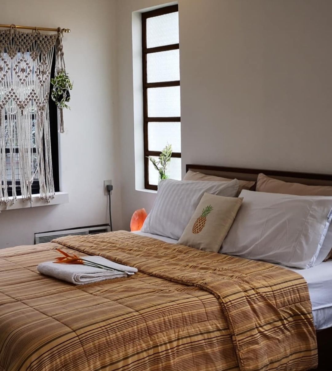 Una cama bien hecha con una colcha escocesa, un tapiz de macramé y una ventana con vista a follaje.