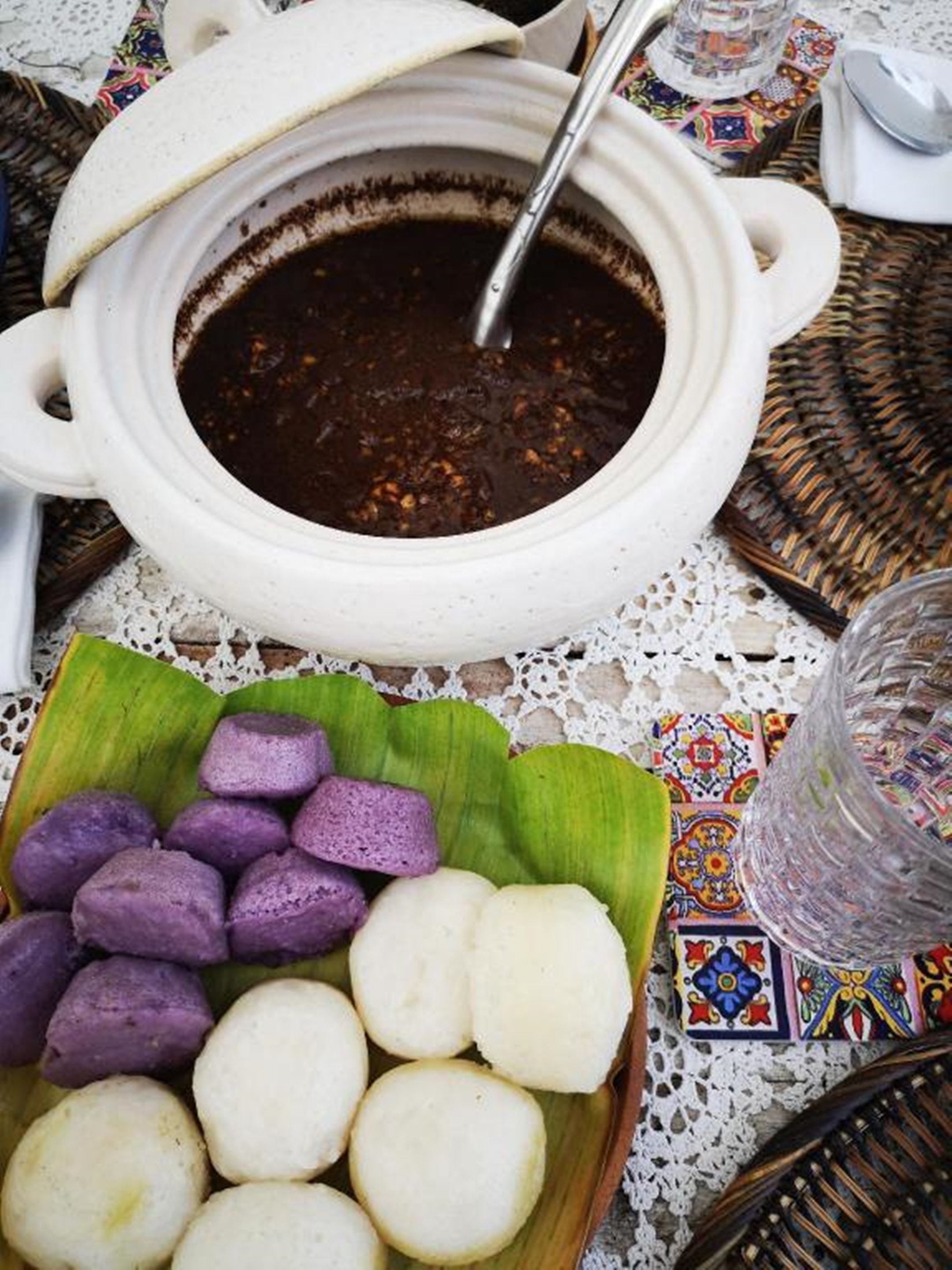 A table with a white ceramic pot containing a dark soup, a bowl of purple and white round food items, and a clear glass.