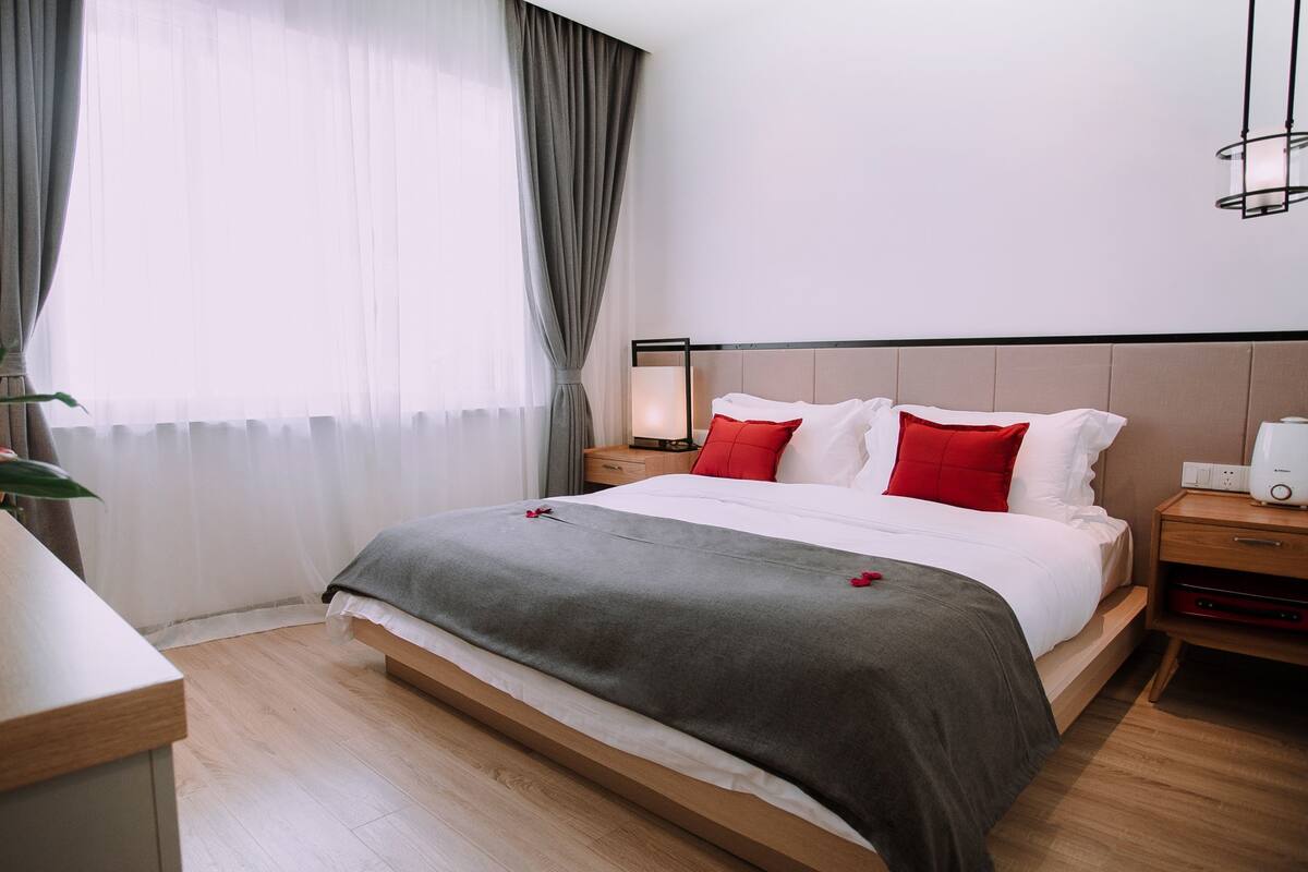 A modern hotel room with a large bed, grey bedding, red pillows, a bedside table with a lamp, and a bedside device.