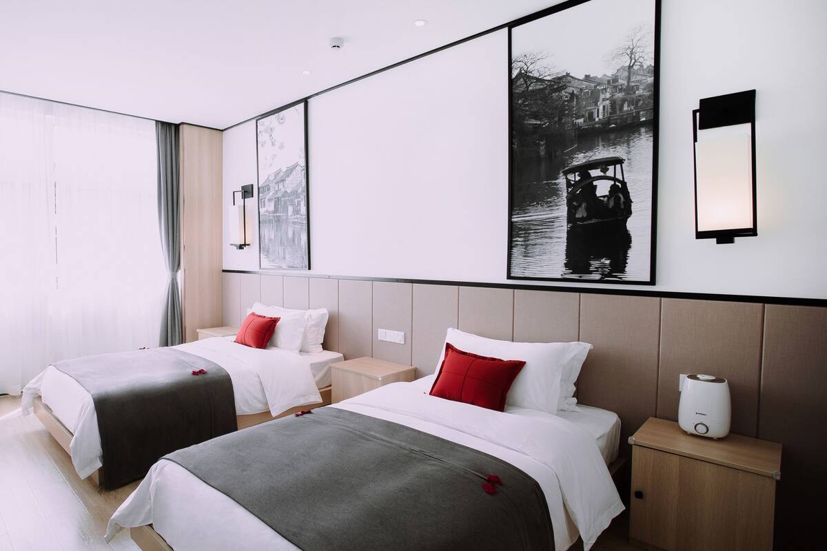 A hotel room with two beds, a large window with curtains, a wall-mounted artwork, and a bedside table with a lamp.