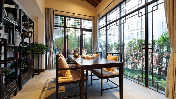 A dining area with a wooden table and chairs, a bookshelf, and large windows with a view of greenery and buildings. - Floral Hotel Wuzhen Ziyemusu (Jiaxing)
