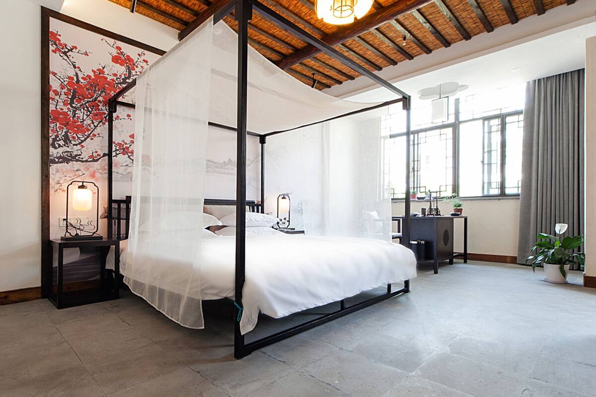 A four-poster bed with a canopy, a nightstand with a lamp, a desk with a chair, and a large window with curtains.
