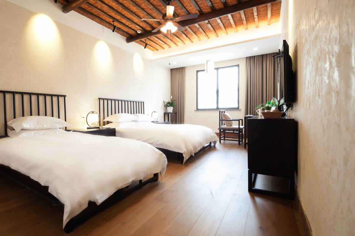 A hotel room with two single beds, wooden flooring, a ceiling fan, a small desk, and a window with curtains.