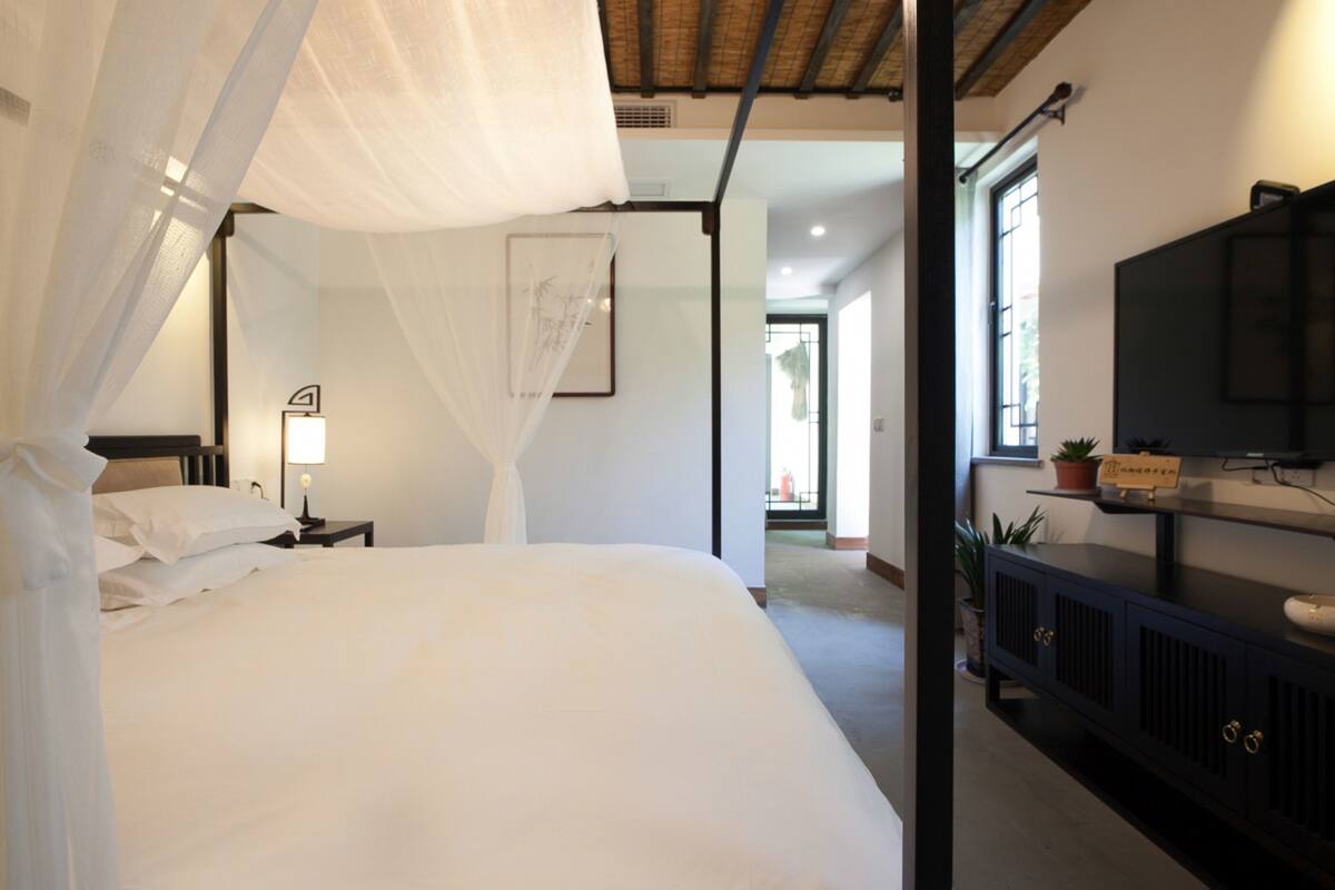 A bedroom with a bed, a nightstand, a bedside lamp, a TV on a cabinet, and a window with a view.