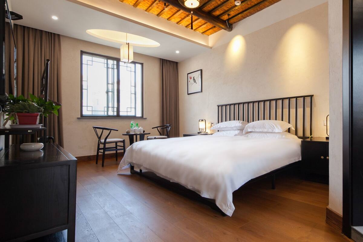 A hotel room with a large bed, a desk with a chair, a small table, and a potted plant.