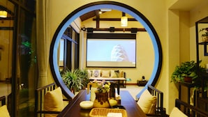 A cozy lounge area with a large circular window, comfortable seating, a coffee table with decorative items, and a projection screen displaying a building image. - Floral Hotel Wuzhen Ziyemusu (Jiaxing)