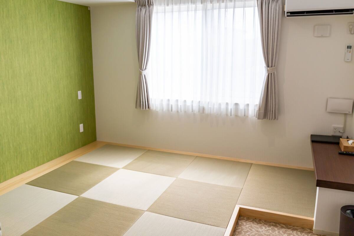 A room with tatami flooring, a green accent wall, a window with curtains, and a desk with a telephone and a box.