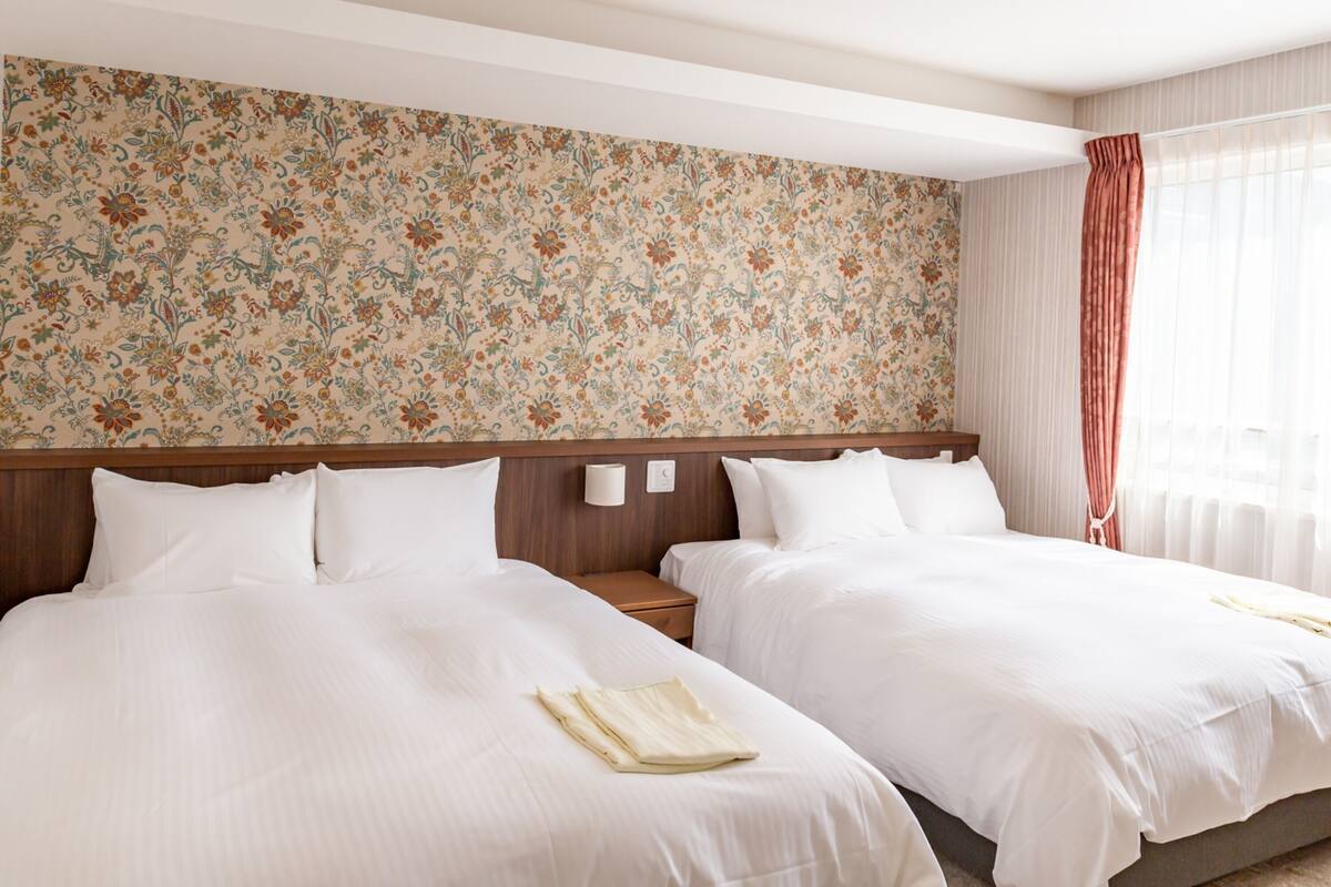 A hotel room with two beds, a floral wallpaper, a wooden headboard, a nightstand, and a window with curtains.