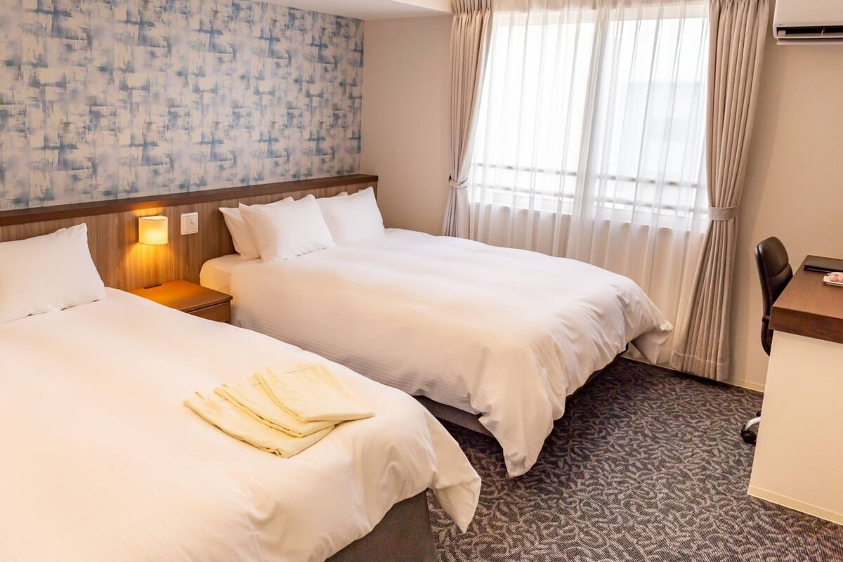 A hotel room with two beds, a desk, and a chair. There is a window with curtains and an air conditioning unit on the wall.