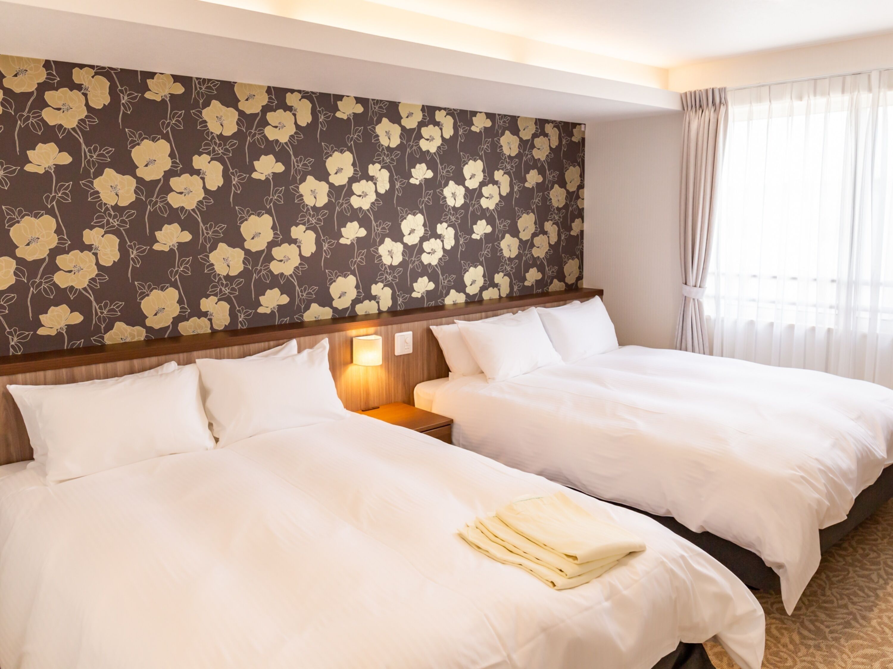 A hotel room with two beds, a floral wallpaper, a nightstand with a lamp, and a window with curtains.