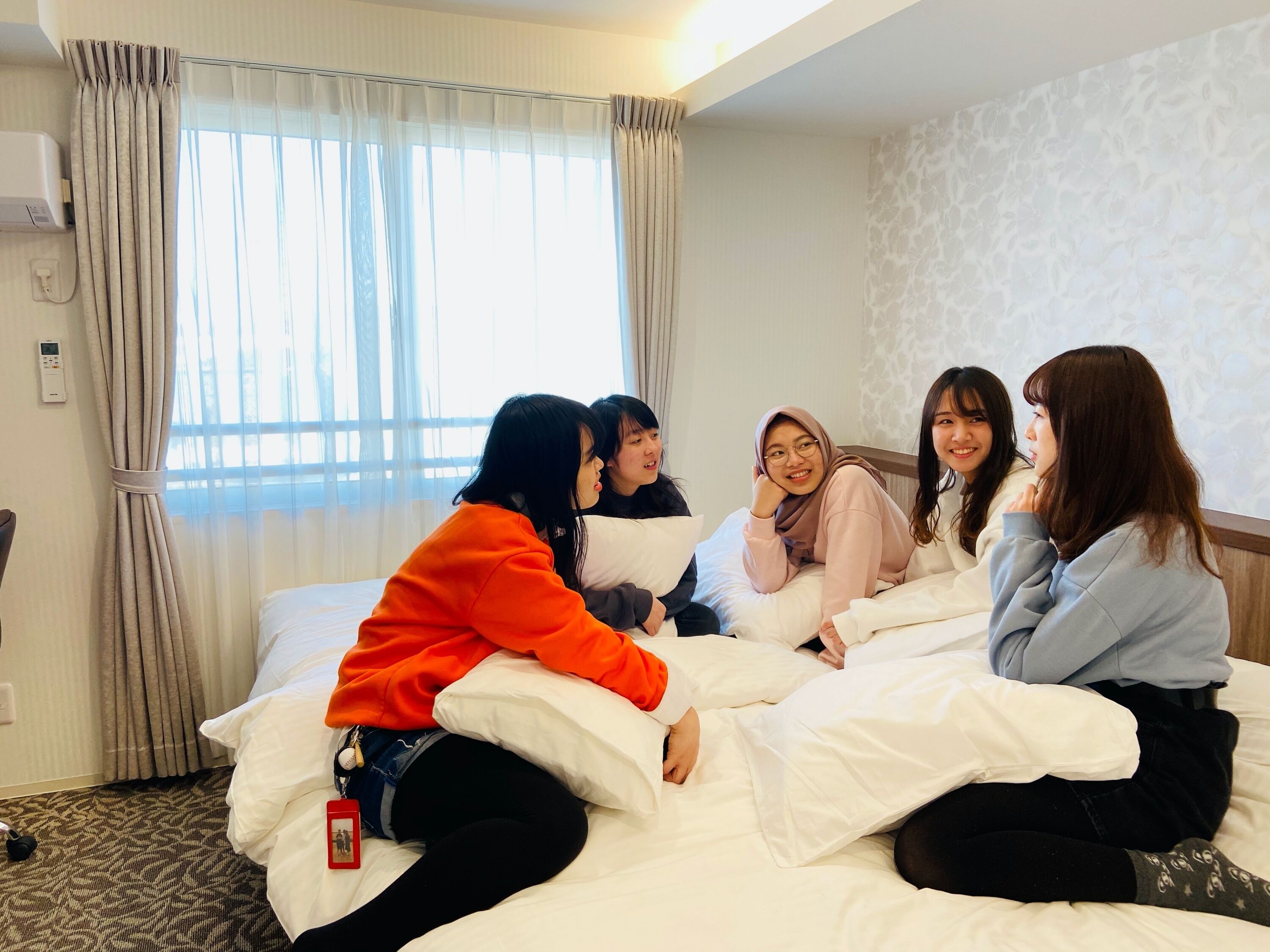 Um grupo de pessoas sentadas na cama de um quarto de hotel, com uma janela, cortinas e um termostato na parede.