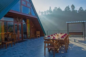 A modern building with large glass windows, a dining area with wooden tables and chairs, and a view of the forested mountains in the background. - Top Mountain Resort (Diyathalawa)