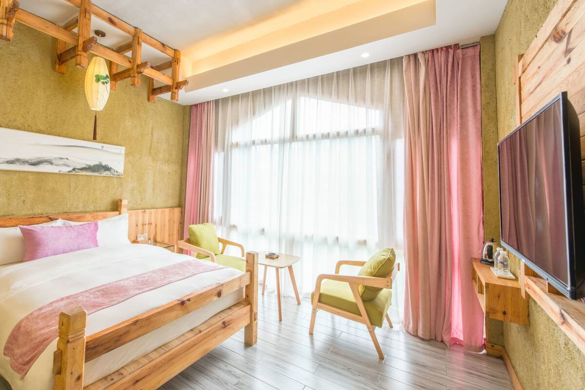 Ein Hotelzimmer mit einem hölzernen Bett, einem kleinen Tisch, zwei Stühlen, einem Fernseher und einem großen Fenster mit Vorhängen.