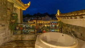 A traditional Chinese-style bathroom with a large bathtub, a balcony with potted plants, and a view of a lit-up town in the background. - Floral Hotel Fenghuang Huercaowu (Xiangxi)