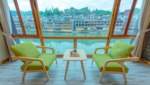 A room with two green cushioned chairs facing a wooden table with a teapot and cups. Large windows offer a view of traditional buildings and a body of water. - Floral Hotel Fenghuang Huercaowu (Xiangxi)
