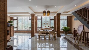A spacious lobby with a wooden reception desk, a seating area with tables and chairs, large windows with geometric patterns, and a staircase. - Floral Hotel Xijiang Banshan Yunfu (Qiandongnan)