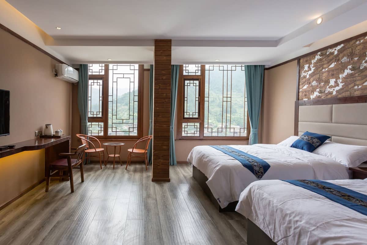 Une chambre d’hôtel équipée d’une télévision, d’un bureau avec une bouilloire, de deux chaises, d’un lit recouvert d’une courtepointe bleue et de grandes fenêtres donnant sur un paysage verdoyant.