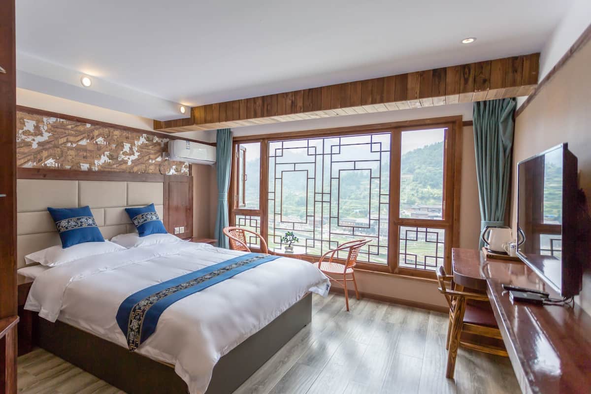 Une chambre à coucher avec un grand lit, une tête de lit en bois, une télévision, un bureau et une fenêtre donnant sur un village.
