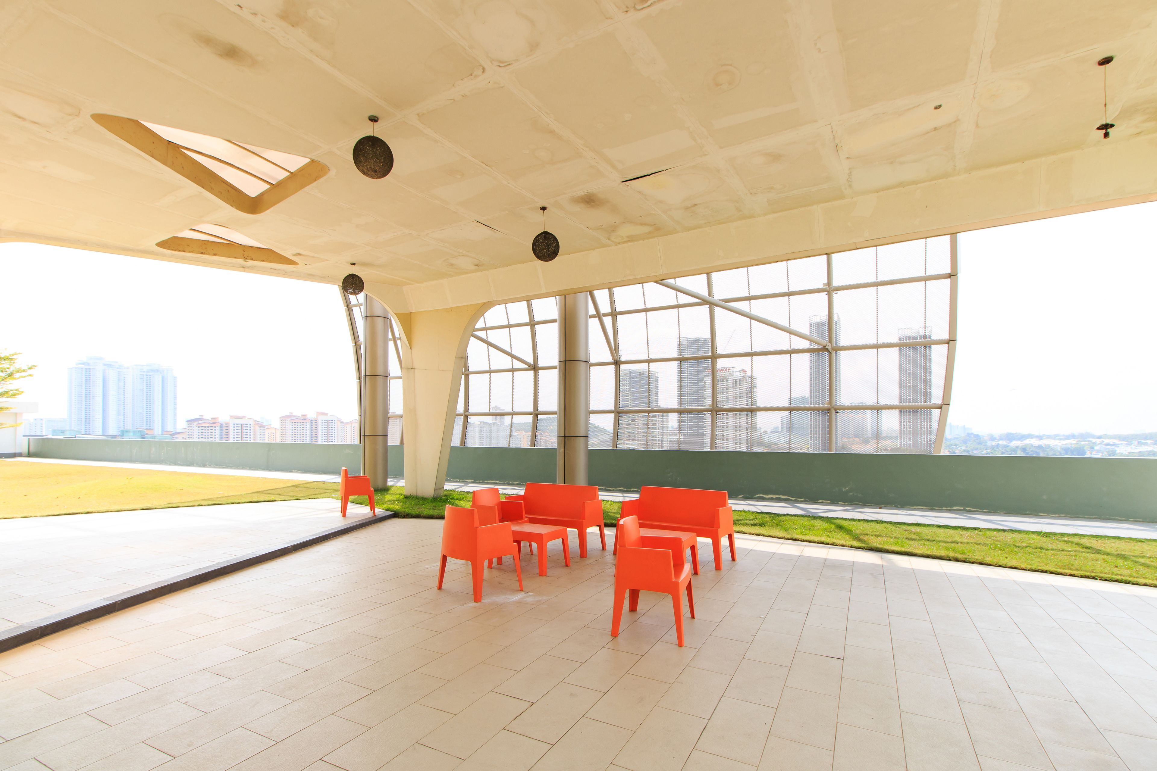 An indoor area with a view of a cityscape through large windows, featuring a tiled floor and orange chairs arranged in a semi-circle.
