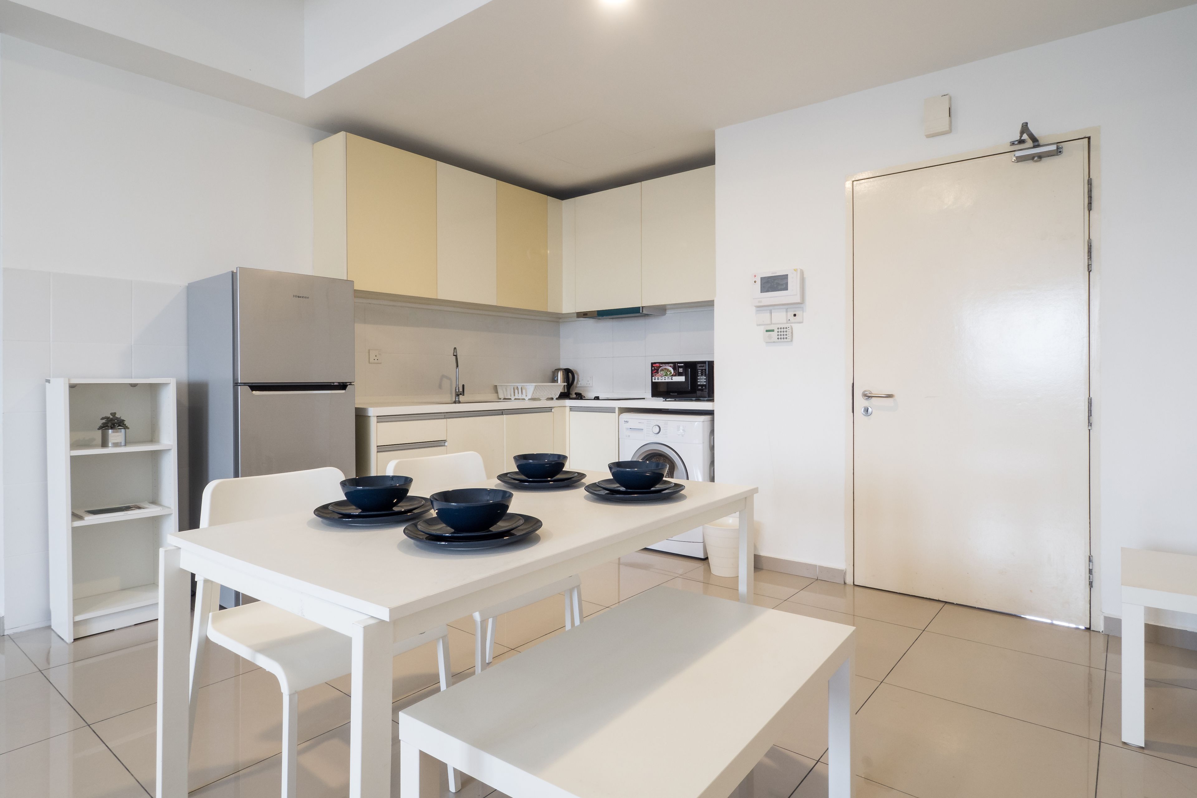 A modern kitchen with white cabinetry, a stainless steel refrigerator, a microwave, and a washing machine. There is a dining table with four bowls and a bench.