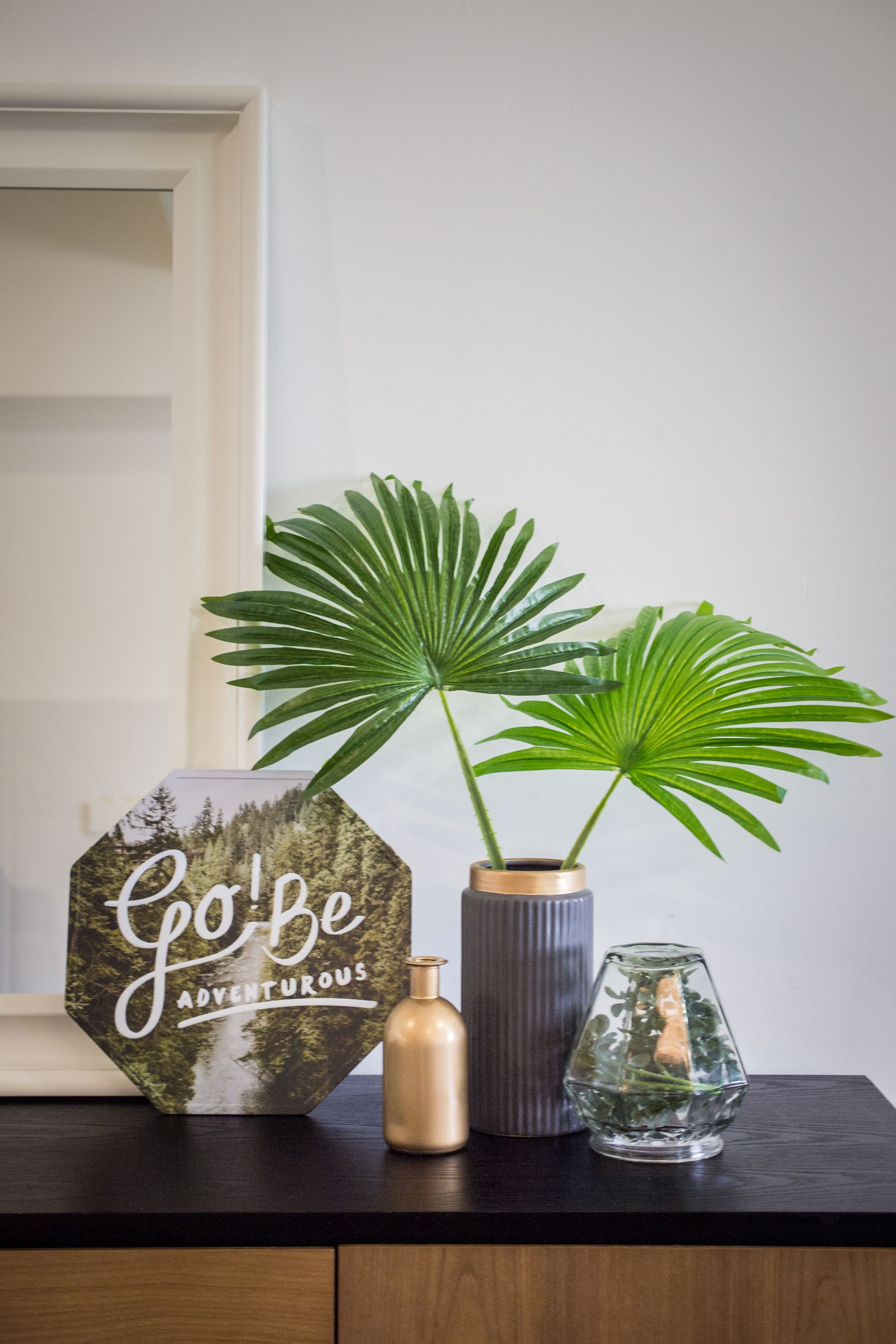 Een houten tafel met een bord waarop staat 'Go be adventurous', een bloempot en een glazen vaas.