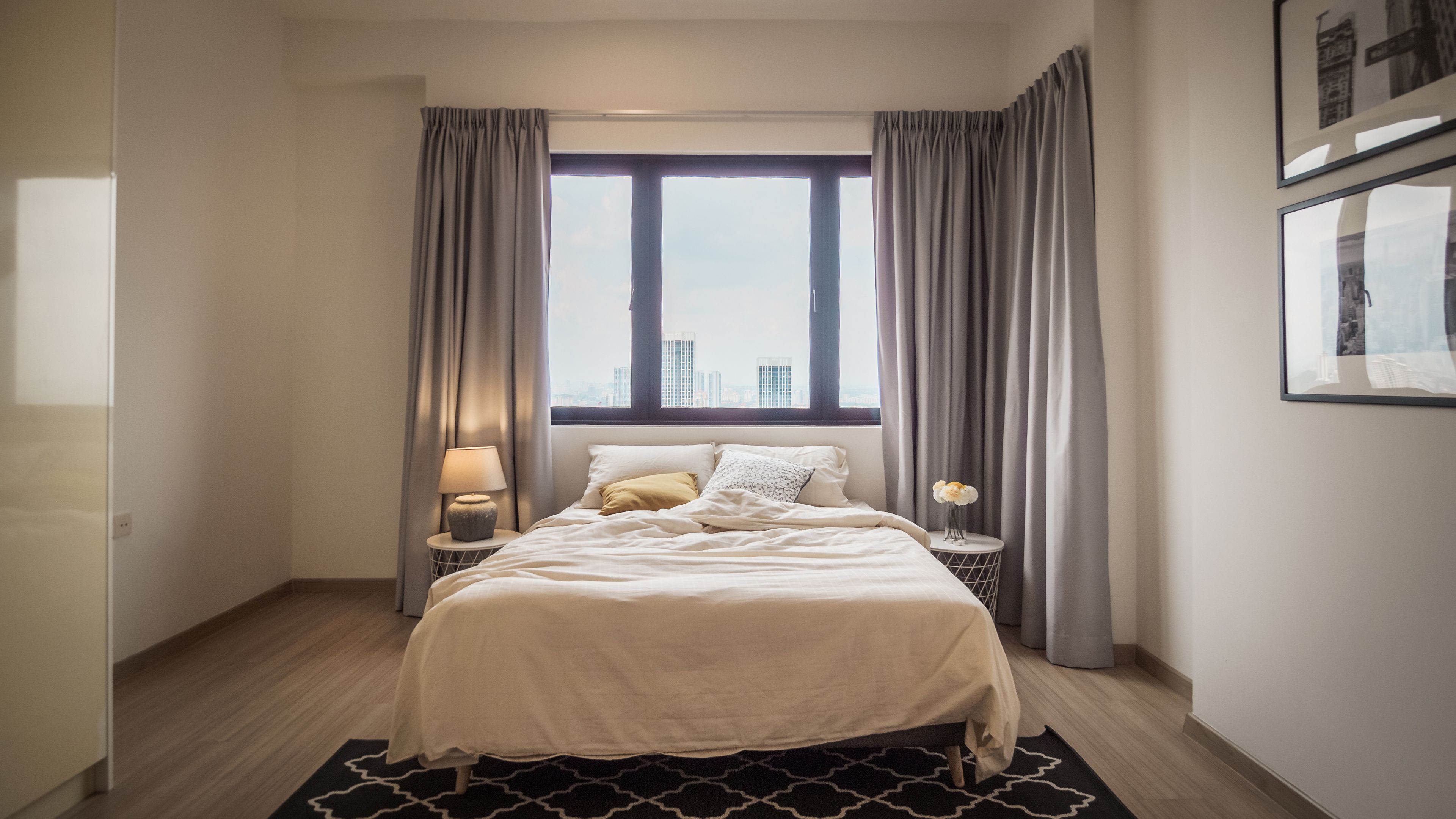 A modern hotel room with a large bed, a nightstand with a lamp, a small table with a vase, and a window with curtains.