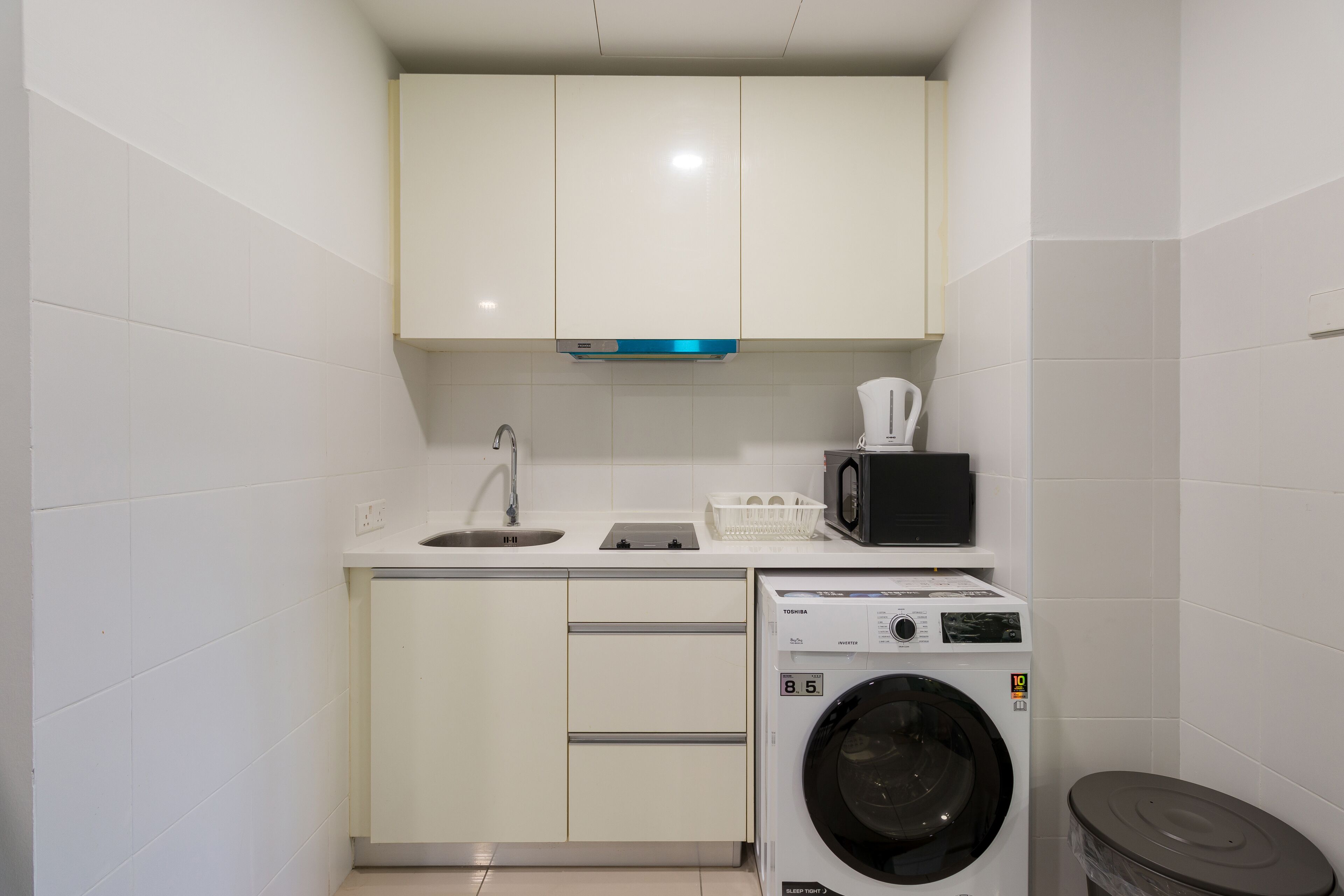 Una cocina compacta con armarios blancos, un lavamanos, un microondas y una lavadora de carga frontal.