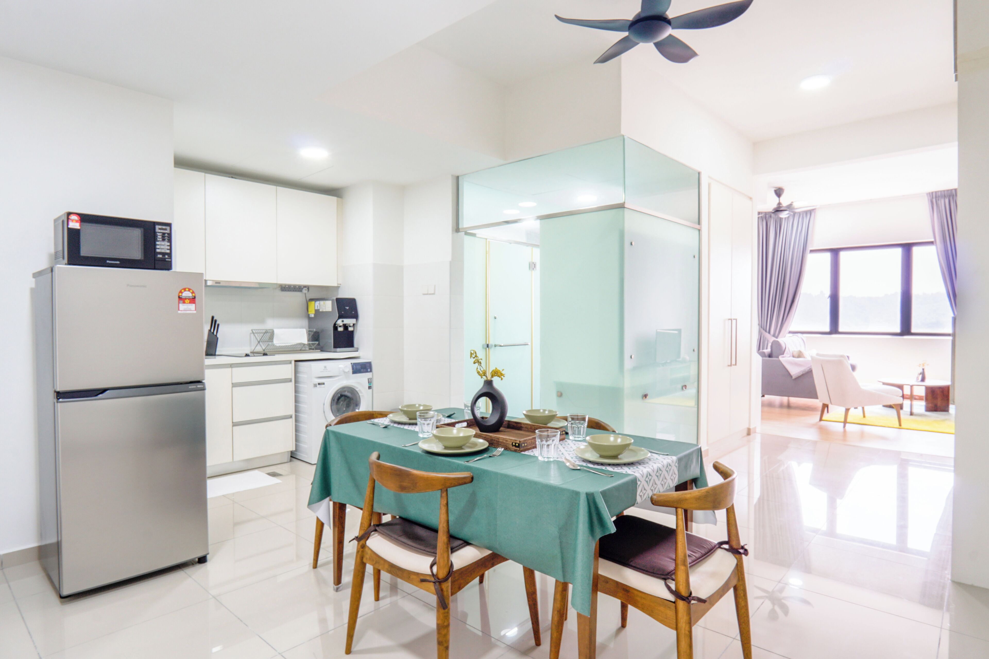 A modern kitchen with a refrigerator, microwave, and washing machine. A dining table with chairs is set for a meal.