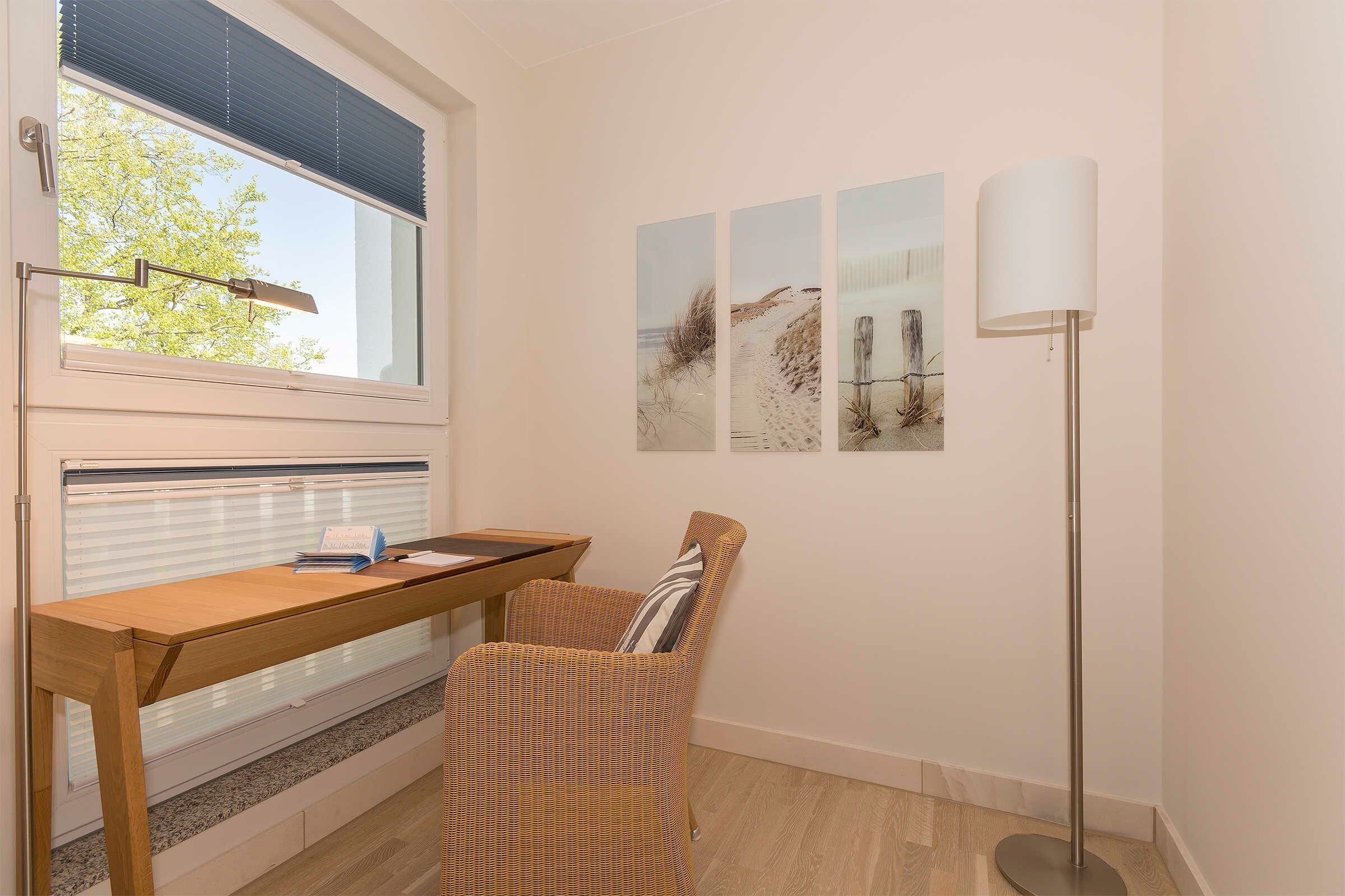A room with a wooden desk, a wicker chair, a standing lamp, and three framed pictures on the wall.