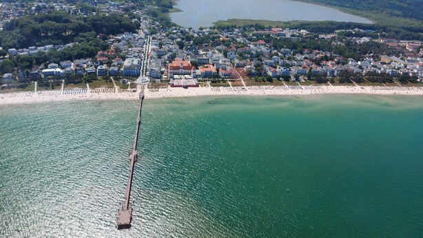 Vue aérienne d’une ville côtière avec une jetée qui s’avance dans l’eau, entourée de bâtiments résidentiels et d’une plage.