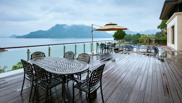 A wooden deck with a table and chairs, overlooking a body of water and mountains.