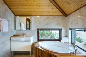 Un baño moderno con techo de madera, una bañera grande, un tocador con espejo y una ventana con vistas a zonas verdes.