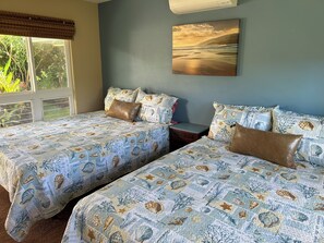 Quarto com duas camas, cada uma com roupa de cama e almofadas combinando, uma janela com vista para vegetação e um quadro na parede.