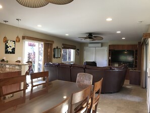 Uma sala de estar ampla com um sofá seccional em formato de L, uma mesa de jantar com cadeiras, uma televisão de tela plana e um ventilador de teto.