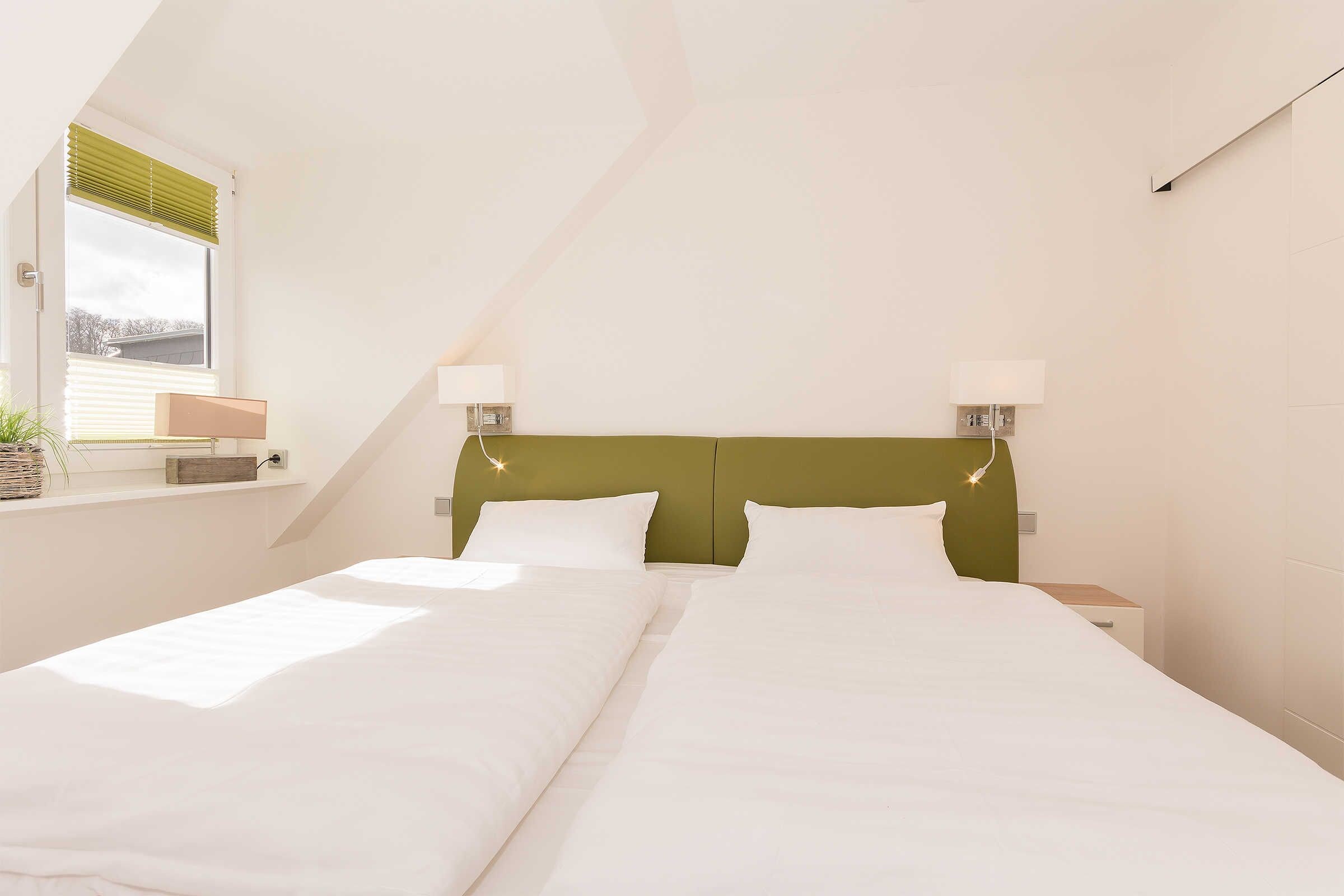 A bedroom with two beds, a green headboard, bedside lamps, a window with blinds, and a small bedside table with a lamp.