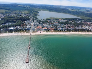 Vista aerea di una cittadina costiera con un molo che si protende nell'acqua, circondata da edifici residenziali e una spiaggia.